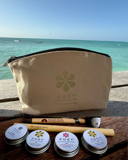 Beige pouch with POSY branding on a wooden surface with a beach in the background. POSY mini bundle of deodorant, shampoo bar, conditioner bar, lotion, lip balm and Nickson travel pen and bamboo straw