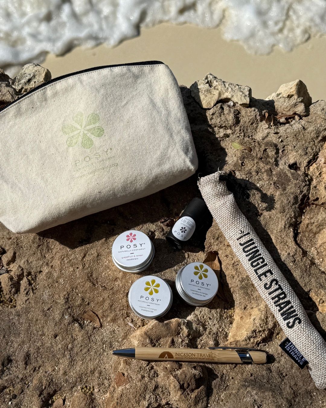 Beige pouch with POSY branding on a wooden surface with a beach in the background. POSY mini bundle of deodorant, shampoo bar, conditioner bar, lotion, lip balm and Nickson travel pen and bamboo straw