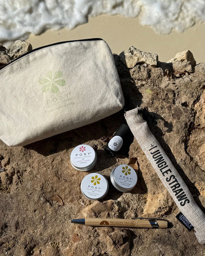 Beige pouch with POSY branding on a wooden surface with a beach in the background. POSY mini bundle of deodorant, shampoo bar, conditioner bar, lotion, lip balm and Nickson travel pen and bamboo straw