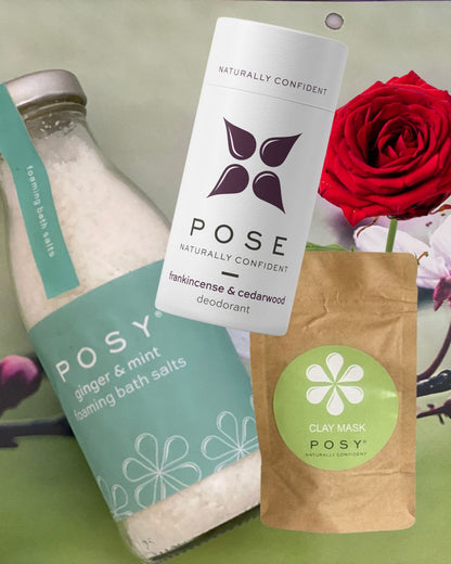 Posy products including a bottle of ginger & mint foaming bath salts, frankincense & cedarwood deodorant and a bag of clay mask on a green background with a red rose.
